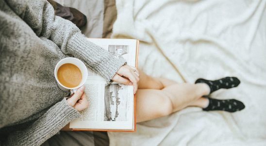 woman holding a cup of coffee at right hand and reading book on her lap while holding it open with her left hand in a well-lit room