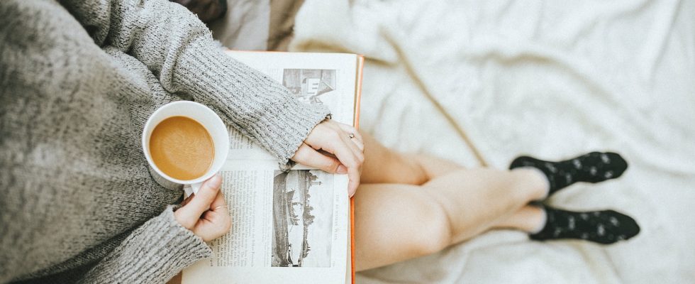 woman holding a cup of coffee at right hand and reading book on her lap while holding it open with her left hand in a well-lit room